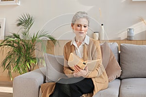 Attractive middle aged woman enjoying reading a book sitting on the sofa in her living room smiling while she reading