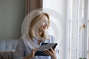 Attractive middle-aged woman holding digital tablet sits on sofa