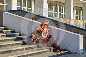 Attractive middle-aged woman wearing stylish coat and shoes sitting with bag on stairs step of office building in early spring at