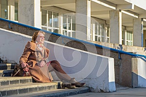 Attractive middle-aged woman wearing stylish coat and shoes sitting with bag on stairs step of office building in early spring at