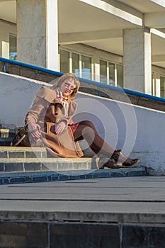 Attractive middle-aged woman wearing stylish coat and shoes sitting with bag on stairs step of office building in early spring at