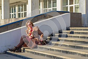 Attractive middle-aged woman wearing stylish coat and shoes sitting with bag on stairs step of office building in early spring at