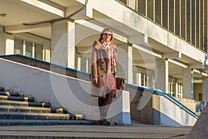 Attractive middle-aged woman wearing stylish coat standing with bag on stairs steps of office building in early spring at sunset.