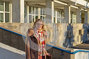 Attractive middle-aged woman wearing stylish coat standing with bag on stairs steps of office building in early spring at sunset.