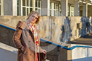 Attractive middle-aged woman wearing stylish coat standing with bag on stairs steps of office building in early spring at sunset.