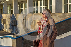 Attractive middle-aged woman wearing stylish coat standing with bag on stairs steps of office building in early spring at sunset.