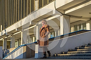 Attractive middle-aged woman wearing stylish coat standing with bag on stairs steps of office building in early spring at sunset.