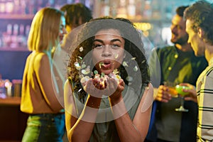 Attractive mixed race young celebrating woman blowing confetti to camera while posing in the night club. Friends