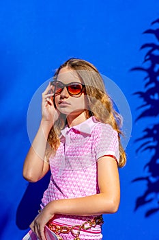 Attractive model Barbie style girl wearing sunglasses, pink tee-shirt and shorts with long hair on blue backdrop