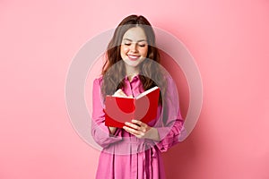 Attractive modern woman writing in her diary, making notes in notebook or planner, looking at her schedule, standing