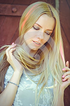 Attractive modest young blond woman playing with hair on red wooden background her hair painted in pink striped striped, in blue d