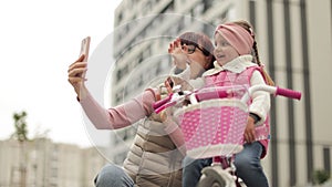 Attractive mother teaching her cute daughter to ride a bicycle, smiling and looking at cell phone