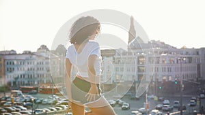 Attractive mulatto young woman with curly hair dancing on a background of modern buildings - sunset