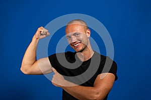 Attractive muscular young man with very short hair and blue eyes pointing his biceps proudly, isolated on blue studio background