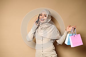 Attractive Muslim Arab woman with beautiful smile in hijab holding color paper bags and talking on mobile phone