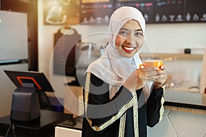 Attractive muslim young asian woman standing at counter in coffee shop, standing and drinking coffee while looking at camera with