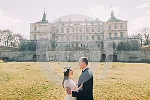 Attractive newlywed couple embracing at green sunny lawn near beautiful ruined baroque palace. Loving groom holding