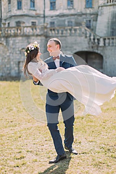 Attractive newlywed couple at green sunny lawn near beautiful ruined baroque palace. Loving groom holding charming bride