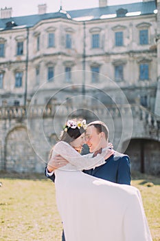 Attractive newlywed pair at green lawn near beautiful ruined baroque palace. Loving groom holding charming bride on his