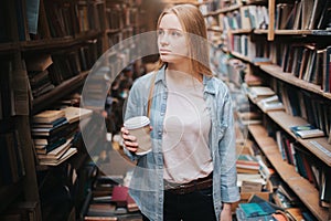 Attractive and nice girl is standing among big and long bookshelfs with old books. She is holding a cup of coffe in her