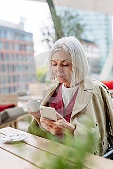 Attractive older woman checking smartphone, drinking coffee at coffee shop. Mature woman spending free time outdoors, in