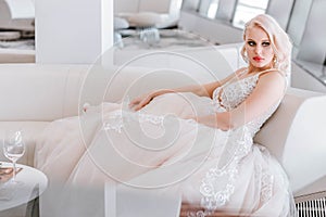An attractive pensive blonde woman with a creative hairstyle for short hair is sitting on a white sofa in the lobby of