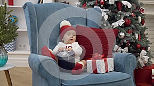 Attractive picture of sweet little baby girl in christmas clothes sitting with new year gifts in comfortable chair near