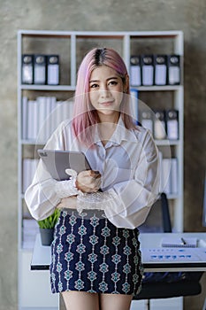 Attractive pink-haired Asian female employee standing in front of a desk Using a touch tablet computer, smiling, emailing business