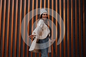 Attractive positive young woman with a beautiful smile in a fashionable hat in a knitted sweater in jeans poses on the background