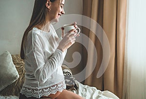 Attractive pregnant woman drinks tea on the bed. Drinking tea looking through a window at home. Last months of pregnancy.