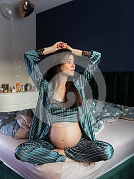 Attractive pregnant woman in pajamas sits in bed and holds her belly.