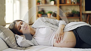 Attractive pregnant woman is resting at home lying on bed and caressing her baby belly with care and love. Loving mother