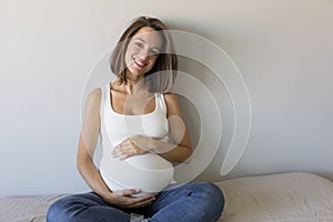 Attractive pregnant woman is sitting in bed, holding her belly and smiling. Last months of pregnancy.Lifestyle indoors