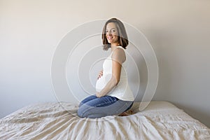 Attractive pregnant woman is sitting in bed, holding her belly and smiling. Last months of pregnancy.Lifestyle indoors