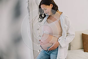 Attractive pregnant woman is standing by the window and holding her belly. Last months of pregnancy