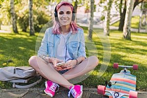 Attractive pretty European beautiful woman with pink hair in headphones listening to music from smart phone sitting in park