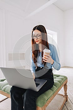 Attractive pretty young woman in eyeglasses freelancer using laptop computer at home, typing text and drinking coffee