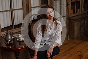 Attractive pretty young woman in a fashionable white shirt in black trendy jeans is sitting on wooden chair in the room. Stylish
