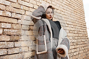 Attractive pretty young woman in trendy checkered coat with white fur in a black stylish t-shirt in vintage gray pants posing