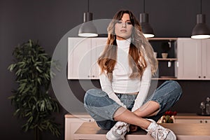 Attractive pretty young woman in a white stylish top in fashionable blue jeans resting on the kitchen vintage wooden table indoors