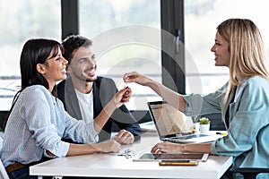 Attractive real-estate agent giving house keys to young couple after signing agreement contract in the office