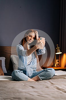 Attractive redhead young woman posing for mobile selfie in bed while sitting on bed at cozy bedroom