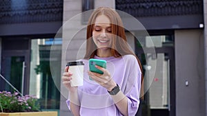 Attractive redhead young woman is using cell and drinking coffee from cup on city street.