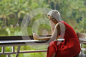 Attractive and relaxed 40s or 50s Asian woman with grey hair and stylish red dress using social media on inernet mobile phone at