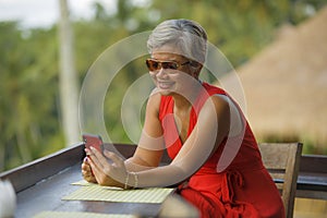 Attractive and relaxed 40s or 50s Asian woman with grey hair and stylish red dress using social media on inernet mobile phone at