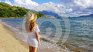 Attractive relaxed young woman in white sundress and straw hat on tropical paradise beach at sunset in Ilhabela, Brazil photo