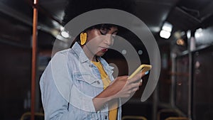 Attractive s,iling young african american woman using smartphone at public transport. Night time. City lights background