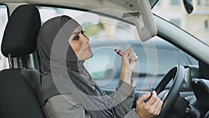 Attractive self-confident Muslim lady applying lipstick sitting in car, coquette