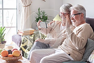 Attractive senior couple relaxing at home on sofa looking at smartphone in video call with family or friends. Smiling senior