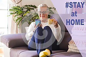 Attractive senior lady sitting on sofa at home using her smart phone for chatting with friends or family drinking a coffee cup -